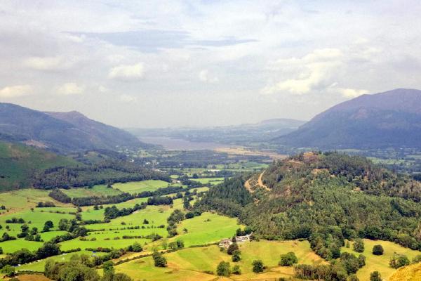 An elevated view over a green valley of patchwork fields in varying shades of green and gold, bordered by dense woodland on the higher slopes. A faint path descends diagonally to the right, and a small white farmhouse sits tucked among the trees. A broad stretch of water is visible on the distant valley floor, with layered fells fading into haze beyond. Overcast sky with soft, diffused light.