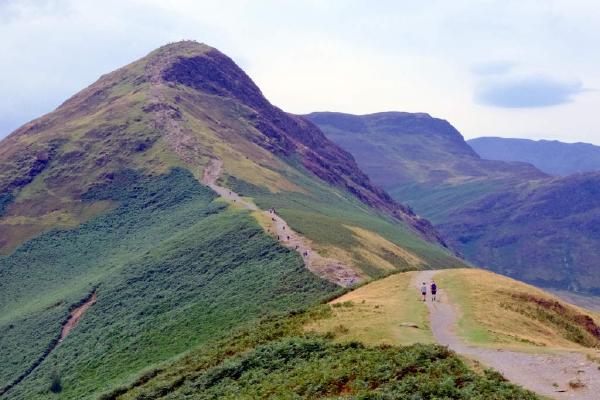 A steep fellside slope covered in a mix of golden-brown grass and dark green vegetation, with a narrow path winding sharply uphill toward a rocky summit. Two small walkers are visible on the path, giving a sense of the gradient. Further fells stack into the distance under a pale, overcast sky.
