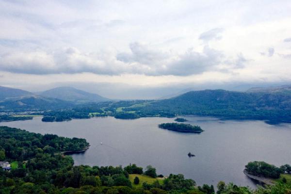A wide view across Derwent Water under an overcast sky, with three prominent wooded islands sitting on the calm, grey-blue surface. The shoreline is densely forested in the foreground, and the town of Keswick is visible along the far shore with its buildings clustered near the water's edge. Layered fells rise behind the town, fading into blue-grey haze under thick cloud cover.