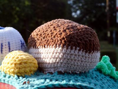A close-up view of crocheted food items sitting on the post box's yarn-bombed surface. The scene includes a white milk bottle with MILK lettered in blue, a brown loaf of bread, a yellow knob of butter and various other crocheted food elements arranged on a turquoise textured base. These detailed food replicas showcase the intricate craftsmanship typical of modern UK postbox and telephone box yarn bombing installations.