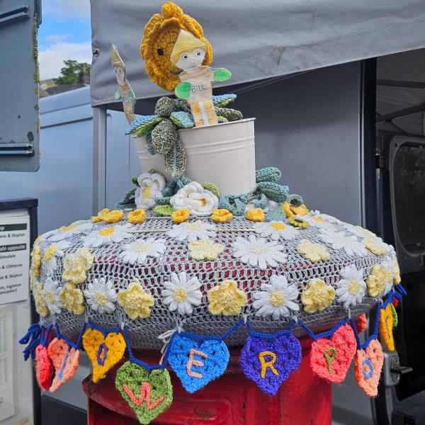 A red pillar postbox in Settle decorated with yarn bombing. On top sits a white crocheted flowerpot containing knitted figures of Bill and Ben the Flowerpot Men alongside Little Weed, the smiling weed character from the classic BBC children&rsquo;s programme. The postbox lid is covered by a grey crochet doily with white and yellow daisies and green foliage. Around the rim hangs a garland of multicoloured crocheted hearts, each embroidered with a letter spelling out FLOWERPOT MEN.