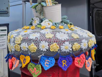 A red pillar postbox in Settle decorated with yarn bombing. On top sits a white crocheted flowerpot containing knitted figures of Bill and Ben the Flowerpot Men alongside Little Weed, the smiling weed character from the classic BBC children's programme. The postbox lid is covered by a grey crochet doily with white and yellow daisies and green foliage. Around the rim hangs a garland of multicoloured crocheted hearts, each embroidered with a letter spelling out FLOWERPOT MEN.