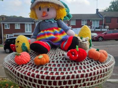 Knitted scarecrow doll in a pompom hat and rainbow skirt, surrounded by knitted corn, apples and pumpkins on a cream crochet base.