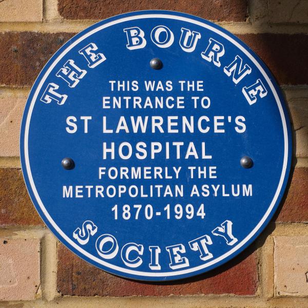 Round blue heritage plaque mounted on a brick wall, reading: The Bourne Society - this was the entrance to St Lawrence&rsquo;s Hospital, formerly the Metropolitan Asylum, 1870-1994.